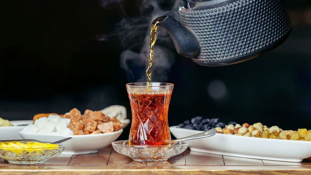 Hot black tea being poured from a cast-iron teapot into an armudu glass, with steam rising above sugar, lemon and dried fruit