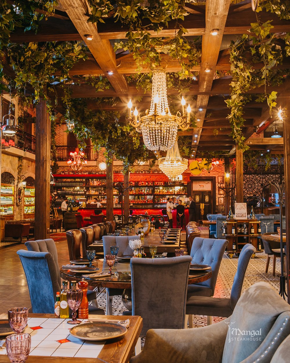The grand dining room at Mangal Steak House in Baku, with crystal chandeliers, velvet seating, exposed wooden beams and trailing greenery above long set tables