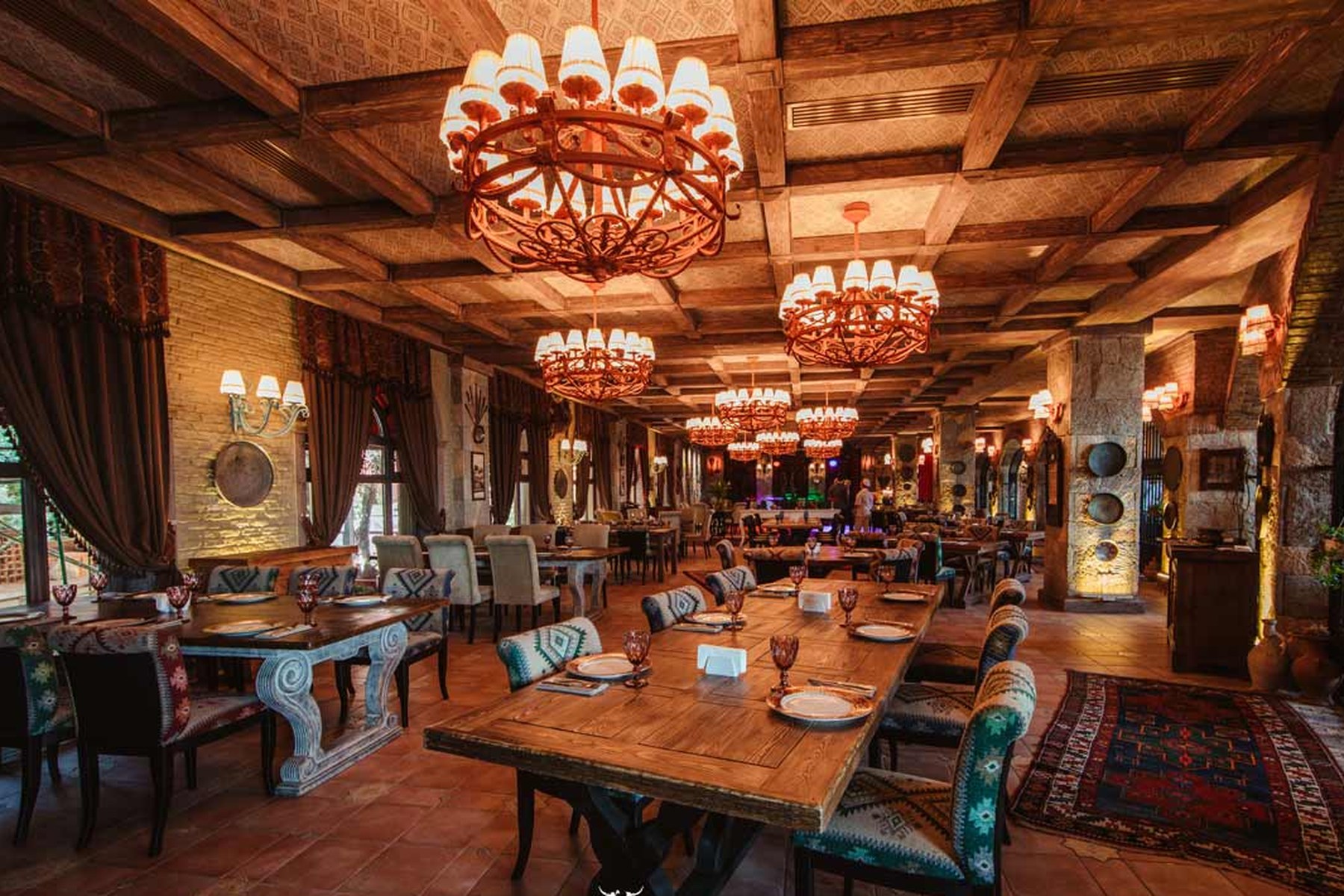 The interior of Mangal Steakhouse Baku, a timbered hall with long tables set beneath wrought-iron chandeliers